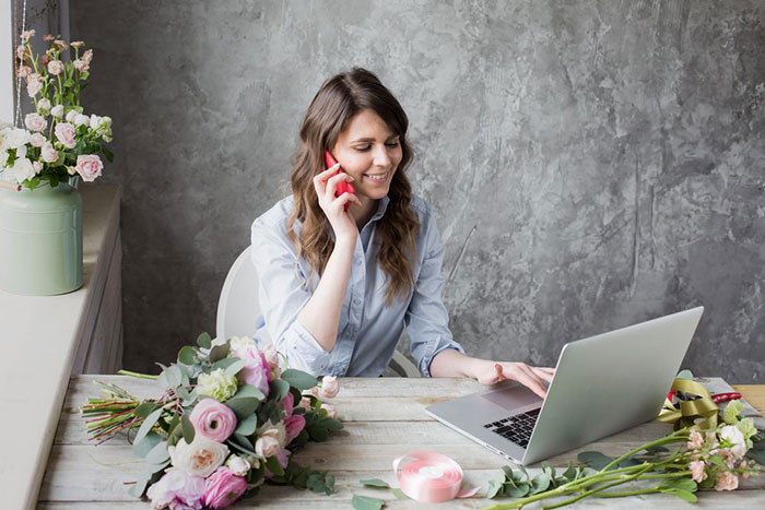 Young florist, on the phone, while working on her laptop, making sure she’s taking action to prevent credit card-not-present fraud.
